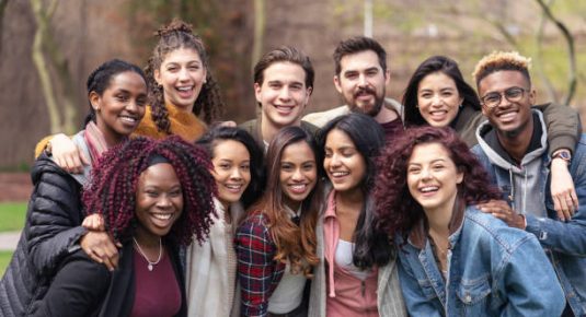 A large sized group of university-aged students, both male and female, are standing out on the campus lawn and smiling into the camera in this educational portrait.They are carefree.