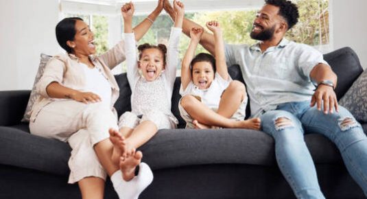 Happy parents and excited children celebrate on the sofa while watching television. Fun family time, relaxing at home and bonding. Mother and father high five, kids cheer for sports team win.