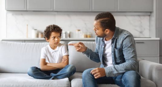 Angry african american father scolding upset little son at home, preteen black boy sitting on couch with arms crossed, dad and male kid having quarrel, suffering misunderstanding, closeup