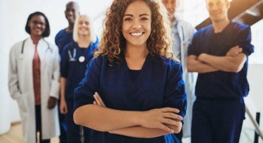 Young female doctor smiling while standing in a hospital corridor with a diverse group of staff in the background